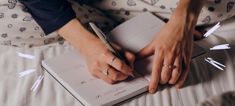 Close up of woman's hands holding a pen and writing in a journal sitting cross-legged on a bed